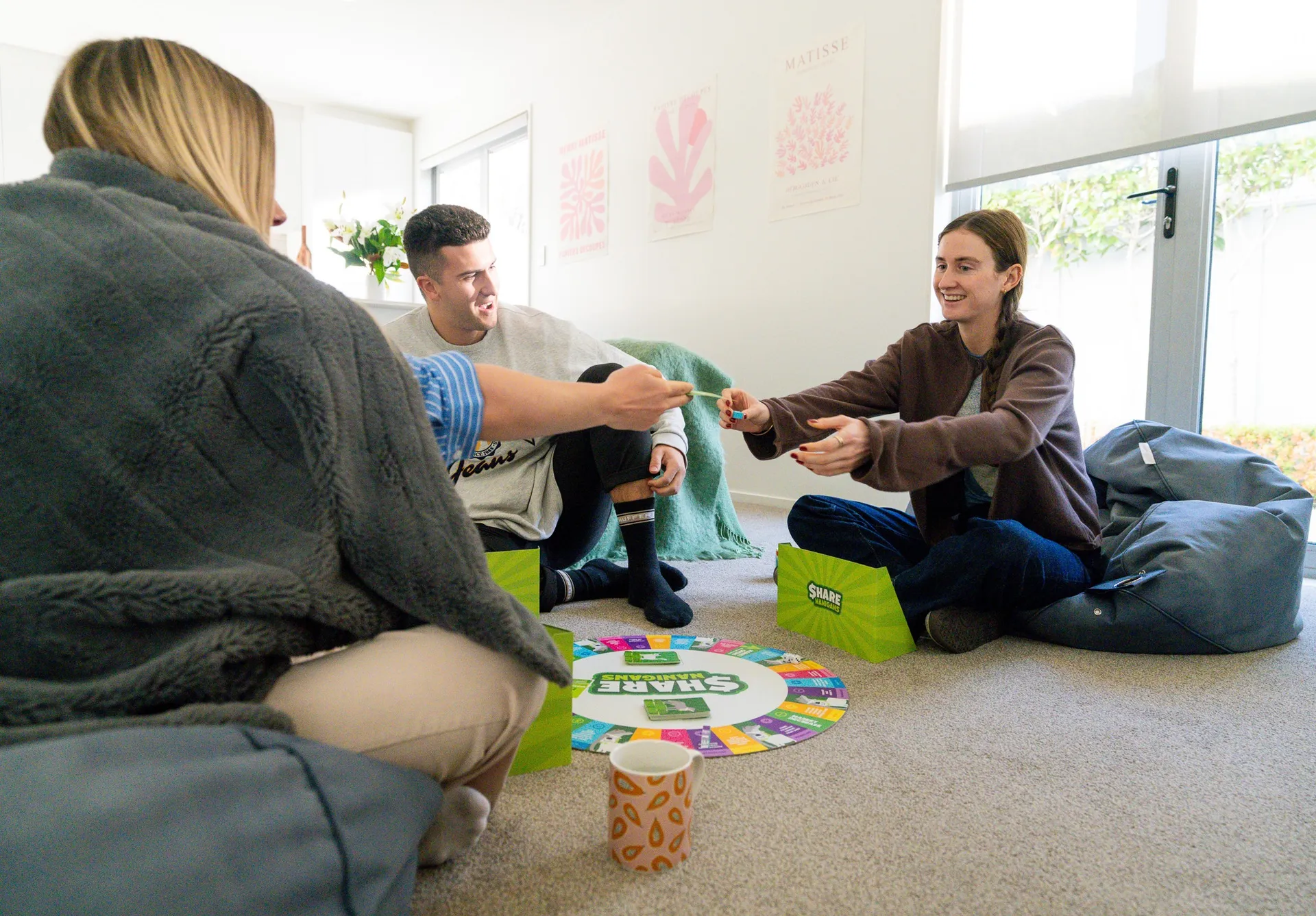 Three friends sitting on the floor of a bright room playing Sharenanigans, the board between them, one passing a card to another