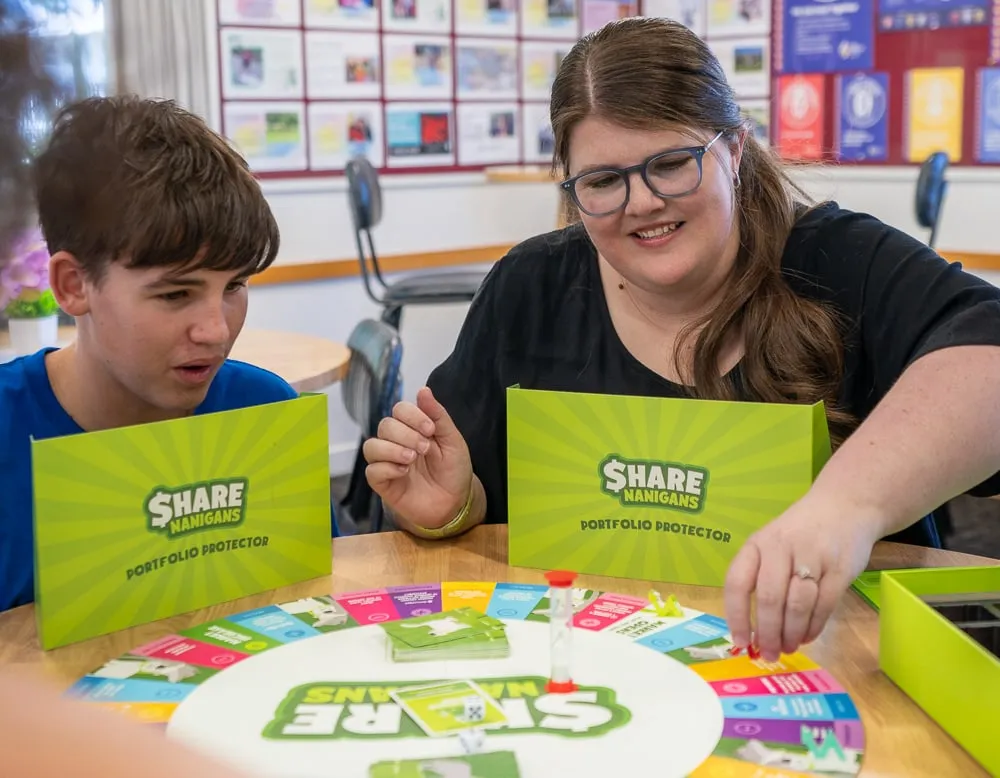 A boy and a woman playing Sharenanigans at a table, behind their Portfolio Protector screens