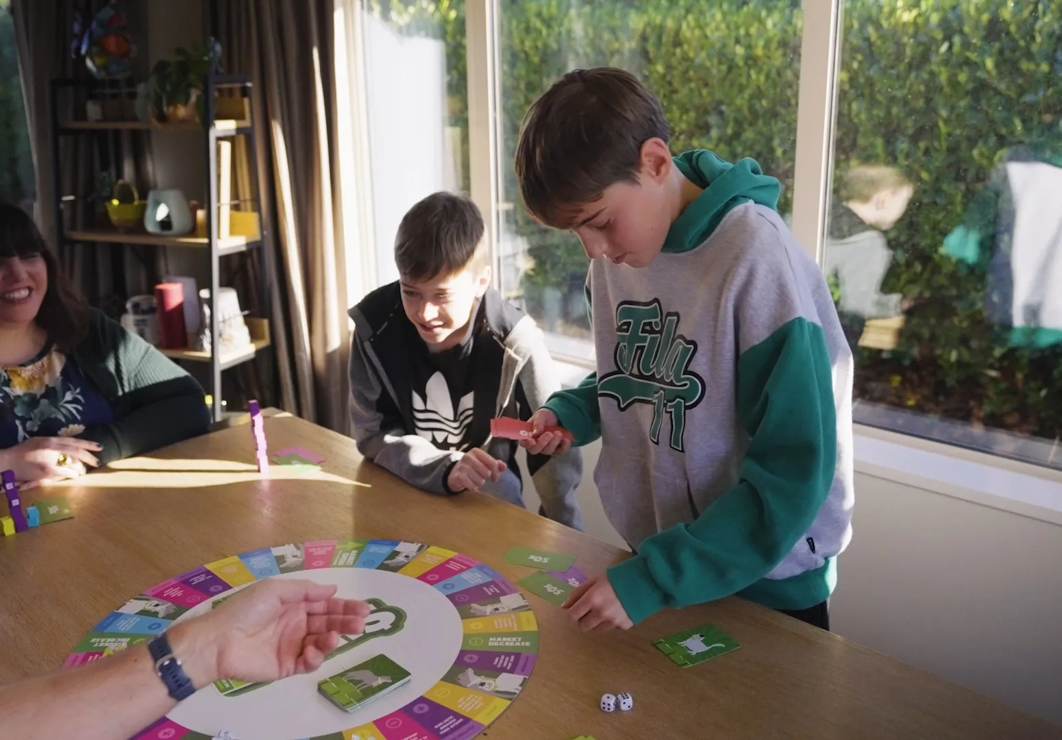 A family playing Sharenanigans at a dining table, two boys leaning over the board while a woman watches and smiles