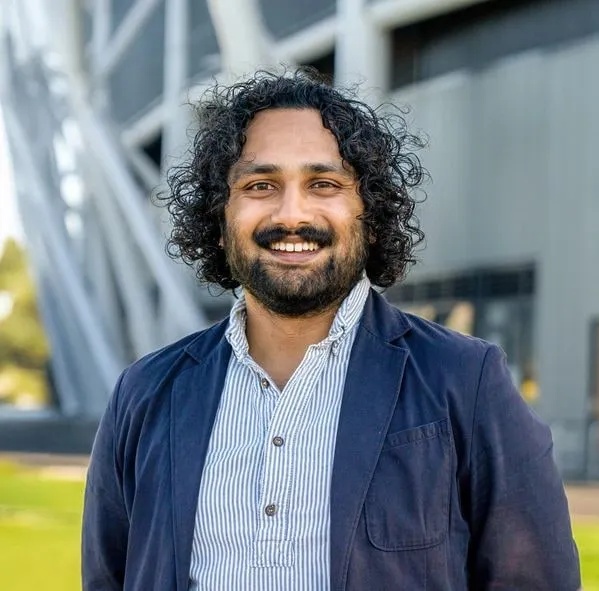 A smiling man with curly dark hair in a navy blazer, outside a modern building