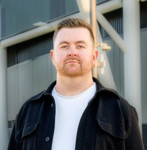 A man with a red beard in a black jacket, outside a modern building