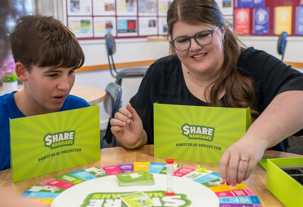 A woman and a boy playing Sharenanigans at a table, behind their Portfolio Protector screens